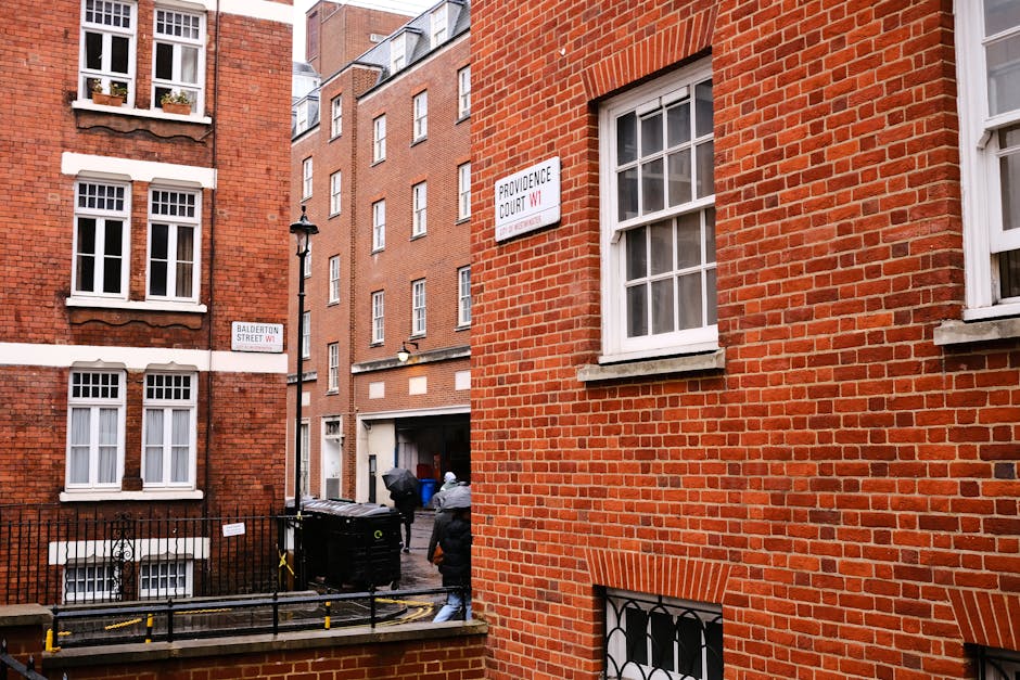 A view of a wet city street in an urban area viewed from a higher floor, with a brick and white facade building on the right side. The street features a small parking area marked with yellow lines and