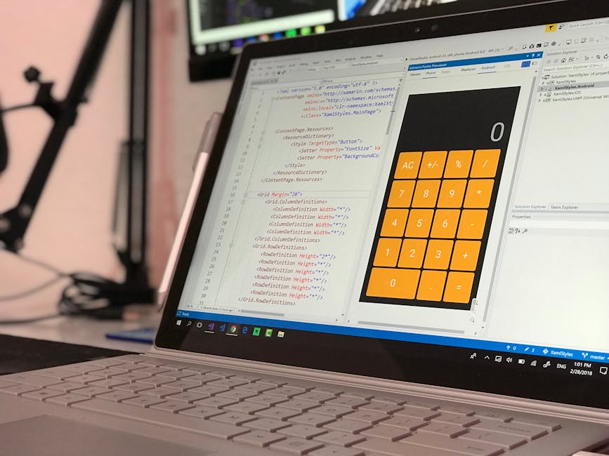 A close-up of a silver laptop on a desk displaying a software development environment with lines of code on the left and a large, orange calculator interface on the right. The calculator features a nu